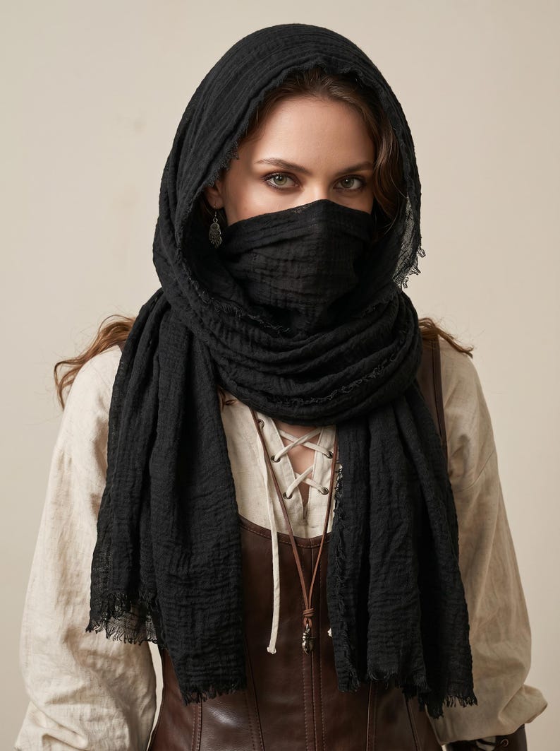 May include: A black textured scarf draped over the head and wrapped around the lower face, featuring frayed edges and a lightweight, crinkled fabric construction. The scarf is worn over a beige long-sleeve shirt and a brown leather-like corset top.