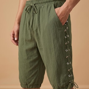 May include: Olive green shorts featuring an elastic waistband with a drawstring, side pockets, and a lace-up detail along the outer leg with silver-toned metal grommets and matching ties at the hem. The fabric has a textured, lightweight appearance.