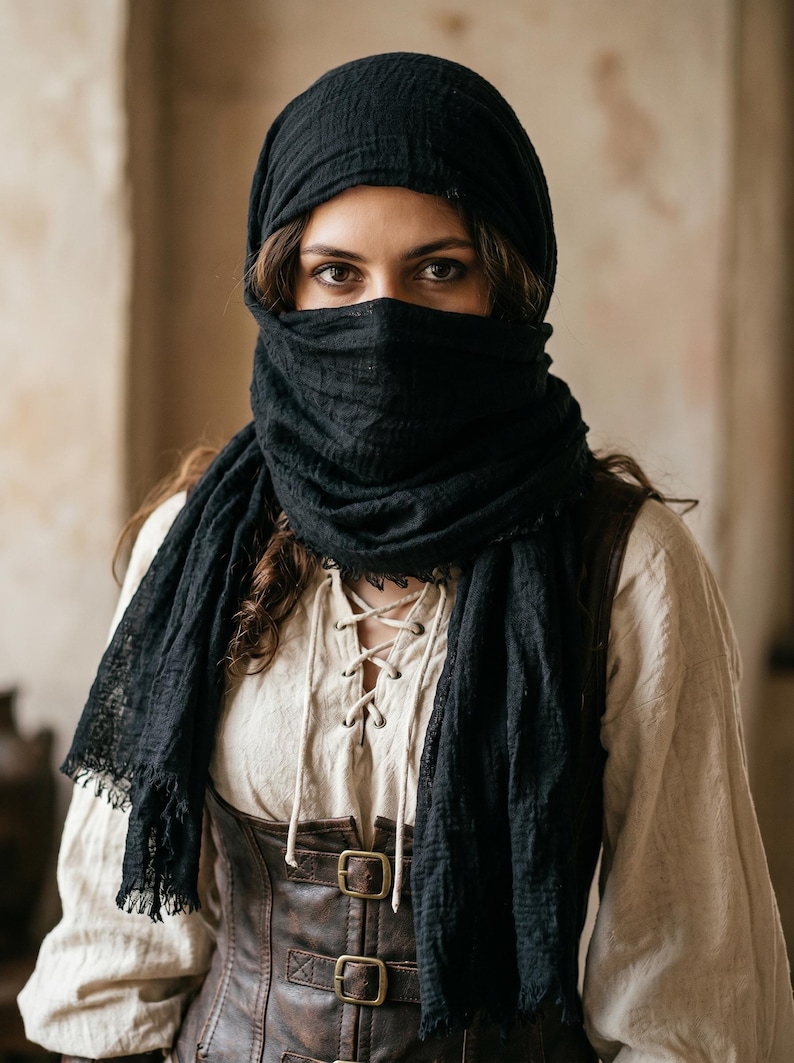May include: A black textured scarf wrapped around the head and face, worn over a cream-coloured long-sleeve shirt with a lace-up front and a dark brown leather-like corset featuring two metal buckles on the front, creating a layered appearance.