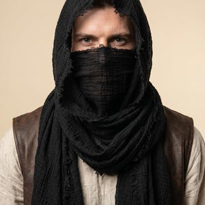 May include: A black, textured, lightweight scarf draped over the head and wrapped around the lower face. The fabric features a crinkled appearance with frayed, raw edges along the borders, creating a layered look over a light-coloured shirt and vest.
