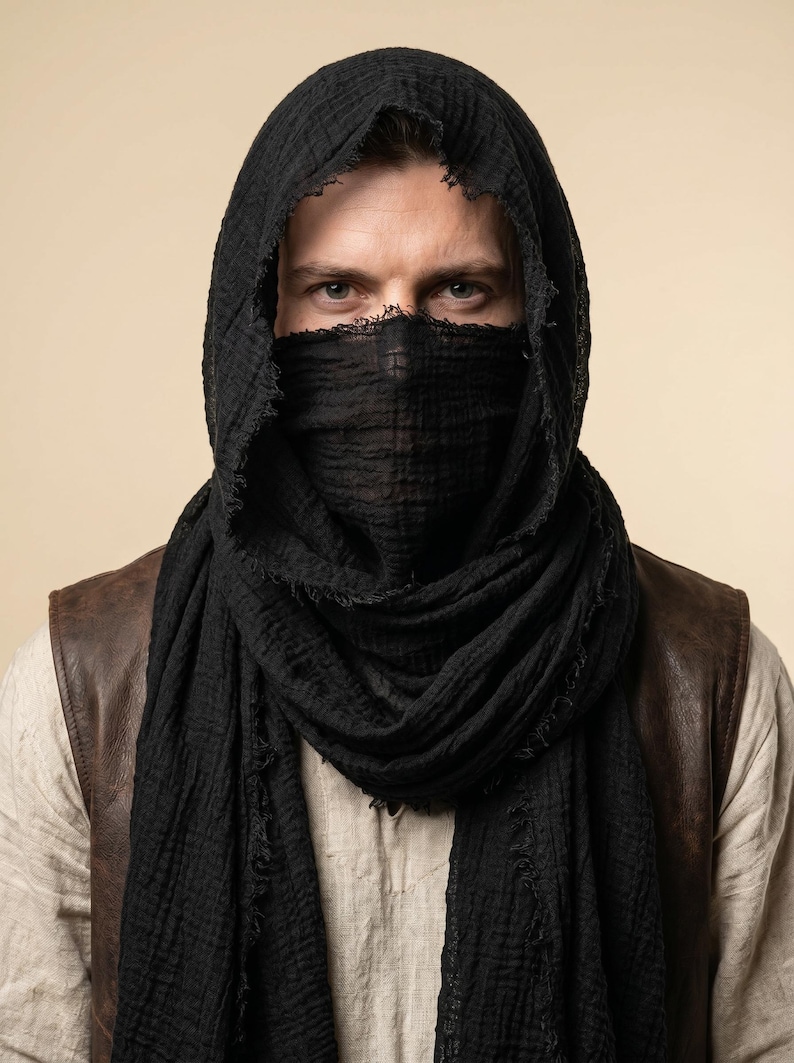 May include: A black, textured, lightweight scarf draped over the head and wrapped around the lower face. The fabric features a crinkled appearance with frayed, raw edges along the borders, creating a layered look over a light-coloured shirt and vest.