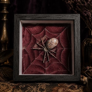 May include: A square black wooden shadow box frame containing a deep red velvet background with white stitched spider web patterns. A spider with long metallic legs and a smooth, speckled, oval-shaped stone body is centered on the web.