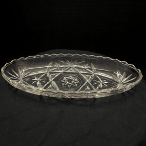 May include: An oval clear glass dish featuring a scalloped edge and a pressed geometric pattern. The design includes multiple starburst motifs and a central star element, creating a faceted appearance across the surface of the shallow tray.