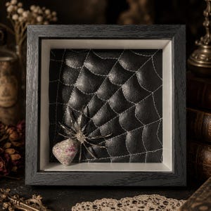May include: A square black wooden shadow box frame containing a black leather-like background with white stitched spider web patterns. A decorative spider with a mottled pink and grey stone body and metallic silver legs is attached to the web.