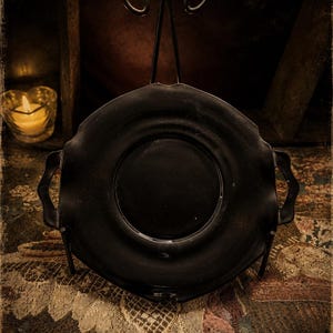 May include: A matte black circular serving tray with a scalloped edge and two side handles, displayed upright on a decorative metal stand. The tray features a smooth, flat center basin and a wide, curved rim, presenting a solid dark finish.