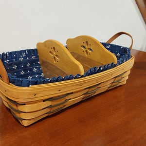 Può includere: Un cesto rettangolare in legno intrecciato con due manici in pelle color cuoio e un rivestimento in tessuto blu scuro con un motivo floreale bianco. Il cesto include due divisori in legno con un design a fiore intagliato, creando tre scomparti.