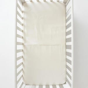 May include: A white wooden crib with vertical slats and a rectangular frame. Inside the crib is a smooth, solid cream-colored mattress cover that fits snugly over the mattress, creating a clean and uniform surface for the sleeping area.