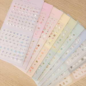 May include: A collection of seven sticker sheets arranged in a fan shape, each featuring a different pastel colour background and various small icons. The top sheet is light purple and labelled Tiny Adore Seal Dream, containing rows of tiny heart and animal stickers.