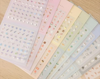 May include: A collection of seven sticker sheets arranged in a fan shape, each featuring a different pastel colour background and various small icons. The top sheet is light purple and labelled Tiny Adore Seal Dream, containing rows of tiny heart and animal stickers.