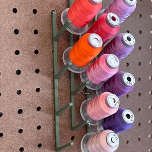 May include: A green plastic thread spool holder with multiple spools of colorful thread. The spools are in shades of red, orange, pink, and purple. The holder is mounted on a brown pegboard.