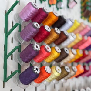 Op de afbeelding: Een verzameling kleurrijke garenspoelen gerangschikt op een wit pegboard. De spoelen zijn in verschillende tinten, waaronder paars, rood, oranje, geel en bruin. Een groene organizer houdt een deel van de spoelen vast. Naai- of knutselthema.