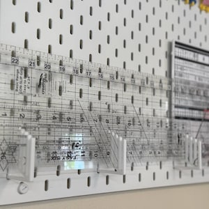 以下が含まれることがあります： 白いペグボード壁面収納には、3つの白いプラスチック製ブラケットが取り付けられており、透明なアクリル製のキルティング定規が数本立てかけられています。定規にはセンチメートルとインチの黒い目盛りが記されており、その中には6インチ×24インチのCreative Grids定規が含まれています。