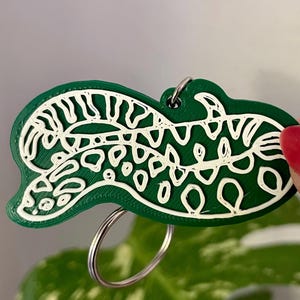 Peut inclure: Un porte-clés vert foncé présentant un motif abstrait en relief blanc, composé de lignes formant une créature serpentine avec des boucles et des points. Le porte-clés est fixé à un anneau métallique argenté pour les clés.