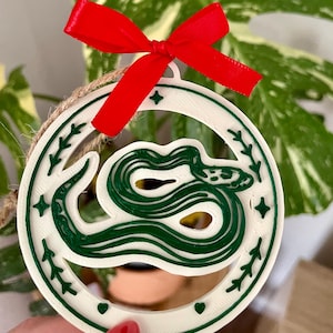 Peut inclure: Une décoration circulaire blanche ornée d'un motif de serpent vert foncé au centre. L'anneau extérieur est décoré de petites feuilles et d'étoiles vertes. Un ruban rouge vif est noué au sommet et une ficelle brune est fixée pour suspendre.