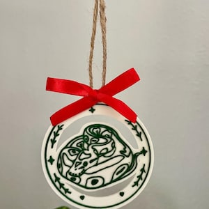 May include: A circular white ornament featuring a green line-art illustration of a coiled snake. The ornament is framed by a border with small leaf and star motifs, topped with a bright red ribbon bow and a natural jute twine loop for hanging.