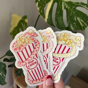 以下が含まれることがあります： 赤と白のストライプのポップコーンバケツに巻き付いたヘビが描かれた3枚のダイカットステッカー。ヘビはピンク、薄いピンク、黄色で、斑点模様があります。各ステッカーには「POP CORN」と「@Velvetscaleexotics」という文字が含まれています。