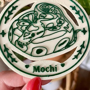 May include: A circular white ornament featuring a raised dark green line art illustration of a coiled snake. The border includes small star and leaf motifs. A small oval plaque at the bottom displays the name Mochi in raised dark green lettering.