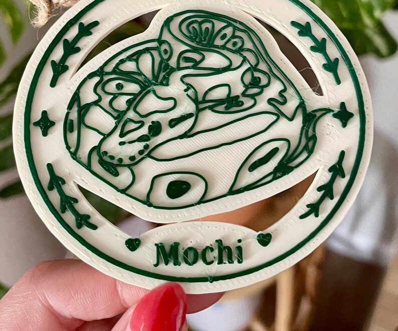 May include: A circular white ornament featuring a raised dark green line art illustration of a coiled snake. The border includes small star and leaf motifs. A small oval plaque at the bottom displays the name Mochi in raised dark green lettering.