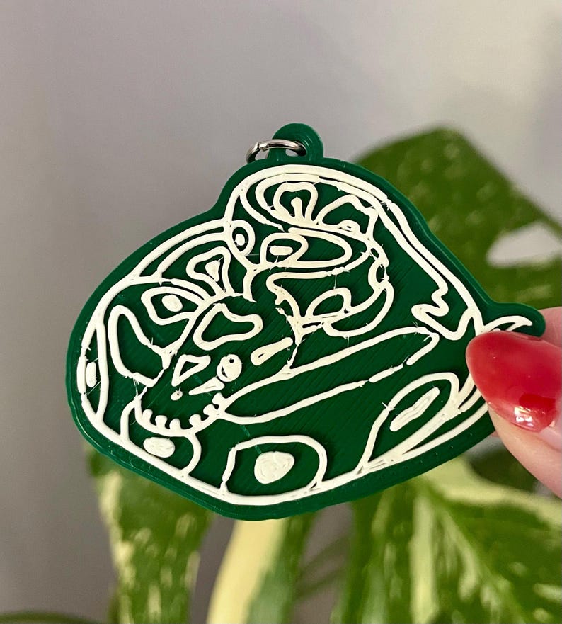 May include: A dark green keychain featuring a white line art design of a coiled snake. The snake is depicted in a continuous, abstract style with visible texture from the 3D printing process. A small metal ring is attached to the top loop.