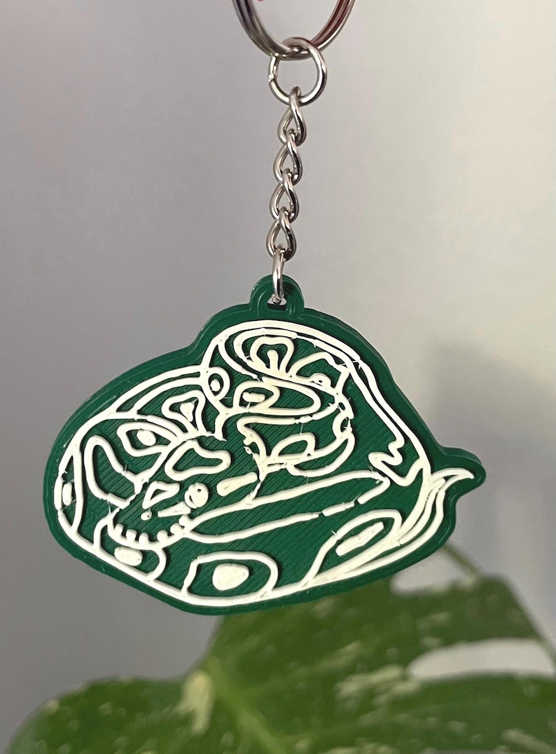 May include: A dark green keychain featuring a white line art illustration of a coiled snake. The pendant is attached to a silver-colored metal split ring by a short chain. The design has a textured surface with raised white lines forming the snake shape.