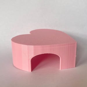 May include: A light pink, heart-shaped object with a smooth, flat top surface and a hollow, arched opening cut into the front side. The structure has vertical, layered sides and a solid, uniform pastel pink color throughout its entire form.