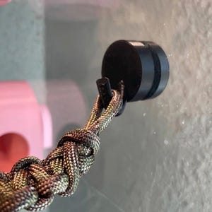 May include: A black circular magnetic mount attached to a surface, featuring a small hook that holds a braided paracord lanyard with a camouflage pattern in shades of olive green, brown, and black, woven into a thick, textured, repeating knot design.