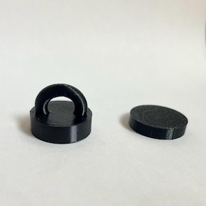 Puede incluir: Dos objetos negros impresos en 3D. Uno es un disco circular con un pequeño lazo arqueado unido a la superficie superior. El segundo objeto es un disco circular negro simple y plano. Ambos artículos presentan una textura en capas visible.
