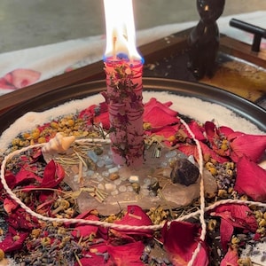 May include: A pink candle with a bright flame, wrapped in twine and dried herbs, stands on a clear crystal base. The candle is surrounded by red rose petals, dried lavender, chamomile flowers, small stones, and a white string on a black plate.