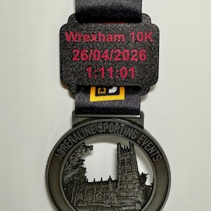 May include: A Wrexham 10K race medal in black and silver. The medal displays "Wrexham 10K", the date "26/04/2026" and a time of 1:11:01. The medal is attached to a black ribbon with "Adrenaline Sporting Events" text.