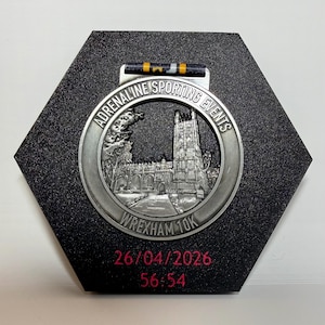 Puede incluir: Medalla plateada de Adrenaline Sporting Events, con una iglesia detallada, montada sobre una placa hexagonal negra. La medalla indica "Wrexham 10K" y muestra la fecha "26/04/2026" y la hora "56:54".