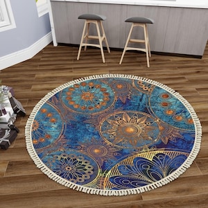 May include: A round area rug featuring a deep blue background with intricate, repeating gold and copper-toned mandala patterns. The rug is finished with a cream-colored fringe border that encircles the entire edge of the circular textile piece.