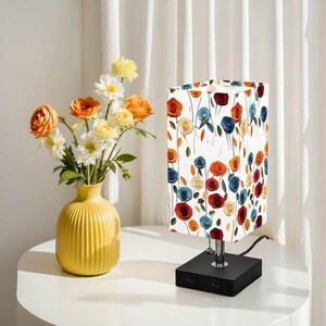 May include: A rectangular table lamp with a floral pattern of red, blue, and orange roses on a white background. The lamp has a black base with USB ports and a chrome stem. A yellow vase with flowers sits nearby.