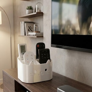 May include: A white cat-shaped remote control holder with several remotes inside. The holder has a cute cat face design and is sitting on a wooden surface. The remotes are black and white.