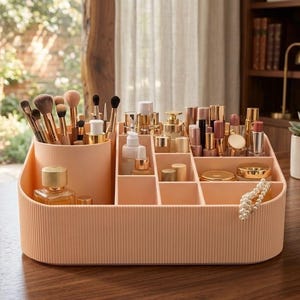 May include: A peach-coloured makeup organiser filled with beauty products. The organiser has compartments and a cylindrical holder for brushes. Lipsticks, perfumes, and compacts are visible, with a string of pearls. Metric measurements are used.