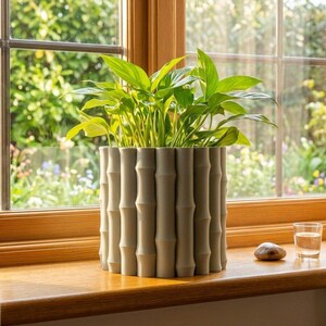 May include: A green leafy plant in a light gray bamboo-style planter sits on a wooden windowsill. A small glass of water and a smooth stone are also on the sill. The window looks out onto a lush green garden.