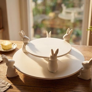 May include: A white, two-tiered serving stand with six bunny figurines supporting the top tier. The stand is made of wood and is set on a wooden table. The background includes a window and a small plate with a yellow item.