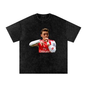 May include: Black t-shirt featuring a graphic of a soccer player in a red and white jersey with the text "Fly Emirates". The player is making a gesture with his hand. The shirt has a vintage, washed look.