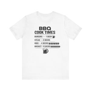 May include: White t-shirt with the text "BBQ COOK TIMES" in black. The shirt lists cooking times for burgers, steak, ribs, and brisket, with beer icons indicating the number of beers needed for each.