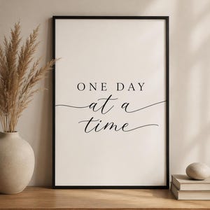 May include: A framed print featuring the phrase "ONE DAY at a time" in black cursive script. The artwork is set against a cream background, enclosed within a black frame. The print is displayed on a wooden surface.