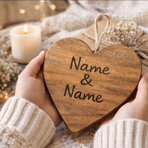 Könnte beinhalten: Ein herzförmiges Holzornament mit der Gravur "Name & Name". Das Ornament wird vor einem verpackten Geschenk und einer brennenden Kerze gehalten, wodurch eine warme Atmosphäre entsteht. Der Hintergrund umfasst Lichter.