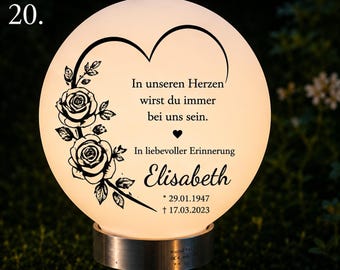 Puede incluir: Una lámpara conmemorativa redonda e iluminada con un gráfico negro de dos rosas y el contorno de un corazón. El texto dice: In unseren Herzen wirst du immer bei uns sein. In liebevoller Erinnerung Elisabeth * 29.01.1947 † 17.03.2023. Se apoya sobre una base de metal.