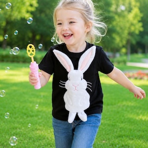 May include: A black short-sleeved t-shirt featuring a large, white, plush rabbit appliqué with pink inner ears and embroidered whiskers, paired with blue denim jeans and a pink plastic bubble wand held in one hand.