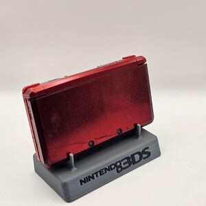 Könnte beinhalten: Eine rote Nintendo 3DS-Handheld-Konsole auf einem grauen 3D-gedruckten Ständer. Auf dem Ständer steht in schwarzer Schrift "NINTENDO 3DS". Die Konsole ist geöffnet und zeigt den Bildschirm.