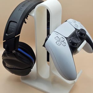 Peut inclure: Support blanc pour casque et manette de jeu, avec un casque noir et bleu et une manette blanche et noire. Le support a un design incurvé. Le casque porte l'inscription "TURTLE BEACH".