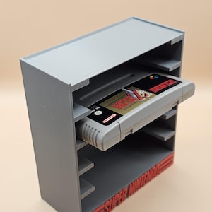 Pode incluir: Um suporte cinza impresso em 3D para cartuchos de jogos Super Nintendo. O suporte tem várias prateleiras e as palavras "SUPER NINTENDO" em vermelho. Um cartucho de jogo Super Nintendo é inserido em uma das prateleiras.