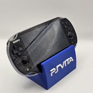 Könnte beinhalten: Eine schwarze Sony PS Vita Handheld-Spielkonsole auf einem blauen Ständer. Der Ständer trägt den weißen Schriftzug "PS VITA". Die Konsole hat eine strukturierte Oberfläche und mehrere Tasten.