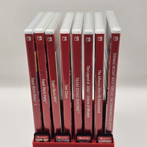 Peut inclure: Un support rouge pour cartouches de jeux avec plusieurs boîtiers de jeux Nintendo Switch. Les boîtiers sont rouges avec du texte blanc, dont "Super Mario Maker 2", "Super Mario Party" et "Teenage Mutant Ninja Turtles". Le support a aussi des emplacements pour les cartouches.