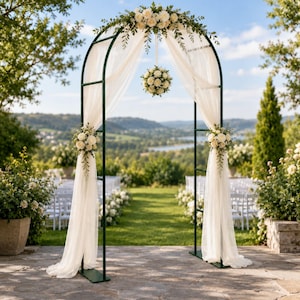 May include: A dark green metal wedding arch draped with sheer white fabric, adorned with clusters of white roses and greenery at the top and sides, and a hanging floral sphere suspended from the center of the arch frame against a landscape.