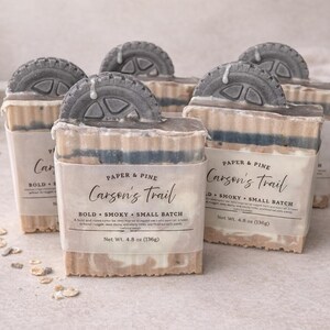 Puede incluir: Varias pastillas de jabón con un diseño en forma de neumático en la parte superior. El jabón tiene un aspecto en capas con tonos beige, azul y blanco. Cada pastilla está etiquetada como "Carson's Trail" con las palabras "Bold, Smoky, Small Batch".