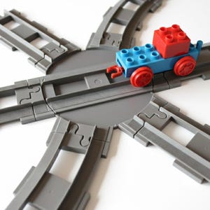 Könnte beinhalten: Ein Spielzeugzug aus blauen und roten Bausteinen steht auf einer grauen Kunststoff-Eisenbahnstrecke. Der Zug hat rote Räder und einen roten Block oben. Die Strecke ist kreisförmig angeordnet.